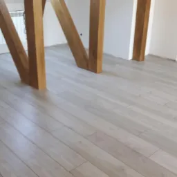 Jasne panele podłogowe ułożone w pomieszczeniu z widocznymi drewnianymi elementami konstrukcyjnymi i grzejnikiem w tle. Na podłodze w oddali widoczny niebieski worek i butelki.