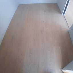 Więcław - Jasne panele podłogowe ułożone w małym pomieszczeniu z widocznymi fragmentami butów na dole kadru.