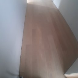 Więcław - Jasne panele podłogowe ułożone w wąskim korytarzu, widok z góry z fragmentem stopy i narzędziem do układania przy ścianie.