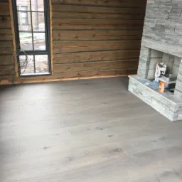 Jasnoszara podłoga z paneli w surowym wnętrzu z drewnianymi ścianami i kamiennym kominkiem w trakcie wykańczania.