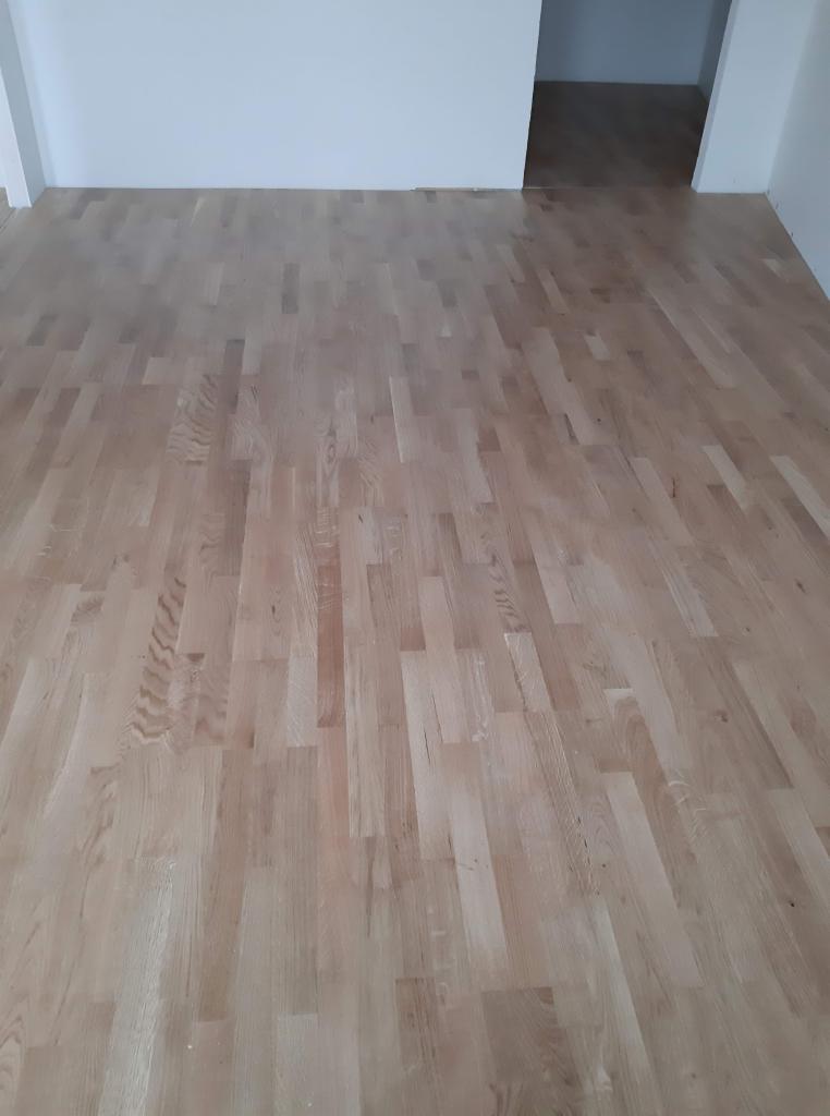 Jasne, drewniane panele podłogowe ułożone w nowym pomieszczeniu z białymi ścianami i otwartym przejściem do innego pokoju.