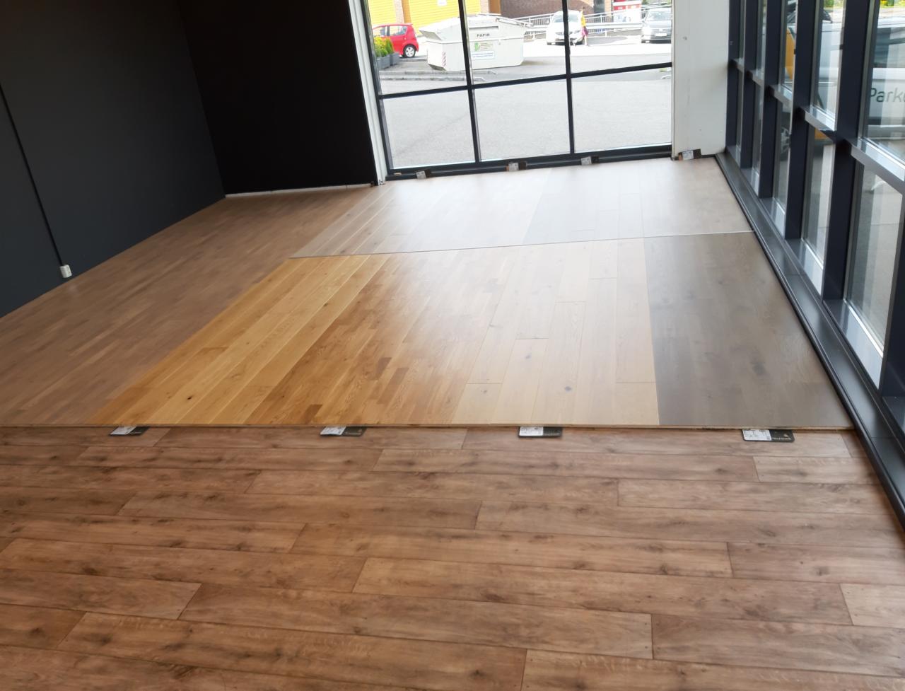 Prezentacja różnych odcieni i rodzajów drewnianych podłóg, z widocznymi próbkami na podłodze, w jasnym wnętrzu z dużymi oknami.