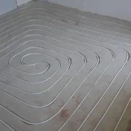Białe rury ogrzewania podłogowego ułożone spiralnie na szarej wylewce betonowej w trakcie instalacji, widoczne gniazdko elektryczne przy ścianie.
