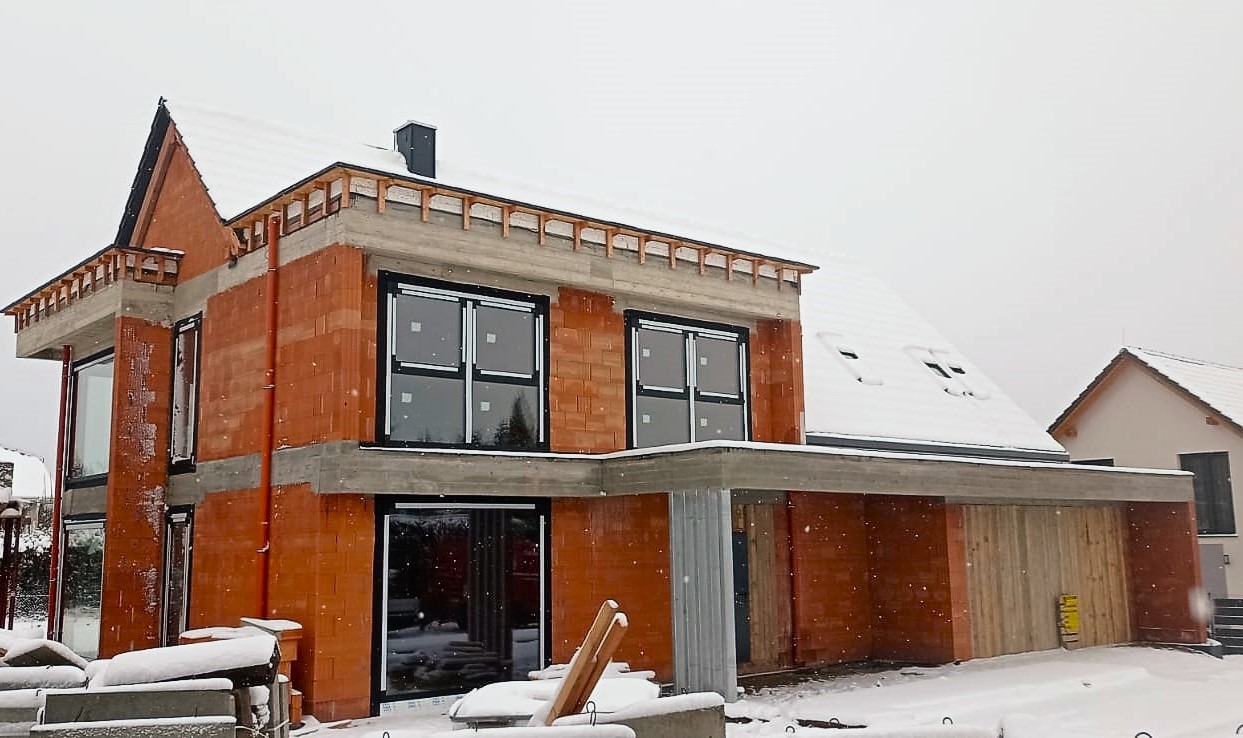 Budynek w trakcie budowy w zimowej scenerii, z zamontowanymi oknami z czarnymi ramami, zabezpieczonymi folią, widoczne bloczki ceramiczne i elementy betonowe konstrukcji.