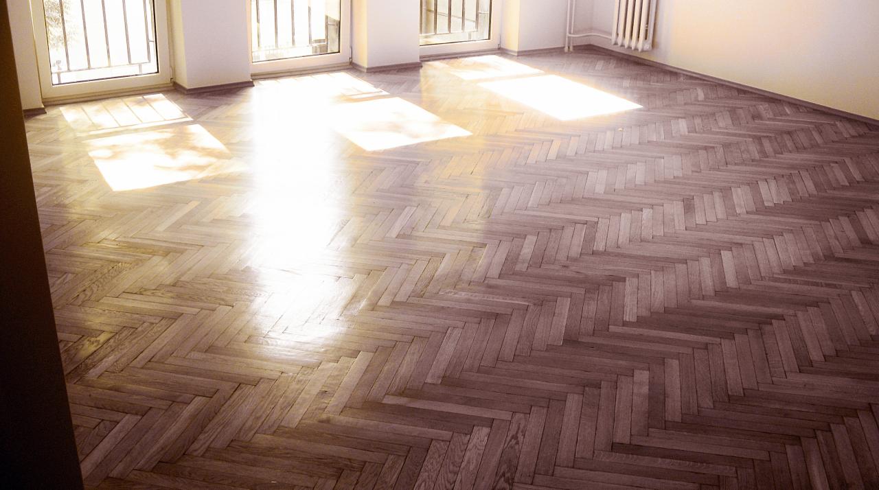 Parkiet ułożony w jodełkę w jasnym, pustym pokoju z promieniami słonecznymi wpadającymi przez okna.