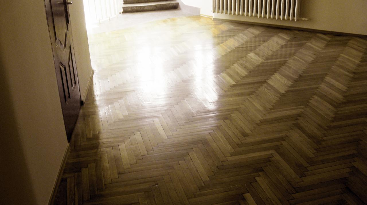 Świeżo wycyklinowana, lakierowana podłoga z parkietu ułożonego w jodełkę, widoczny fragment drzwi i schodów, ciepłe oświetlenie.