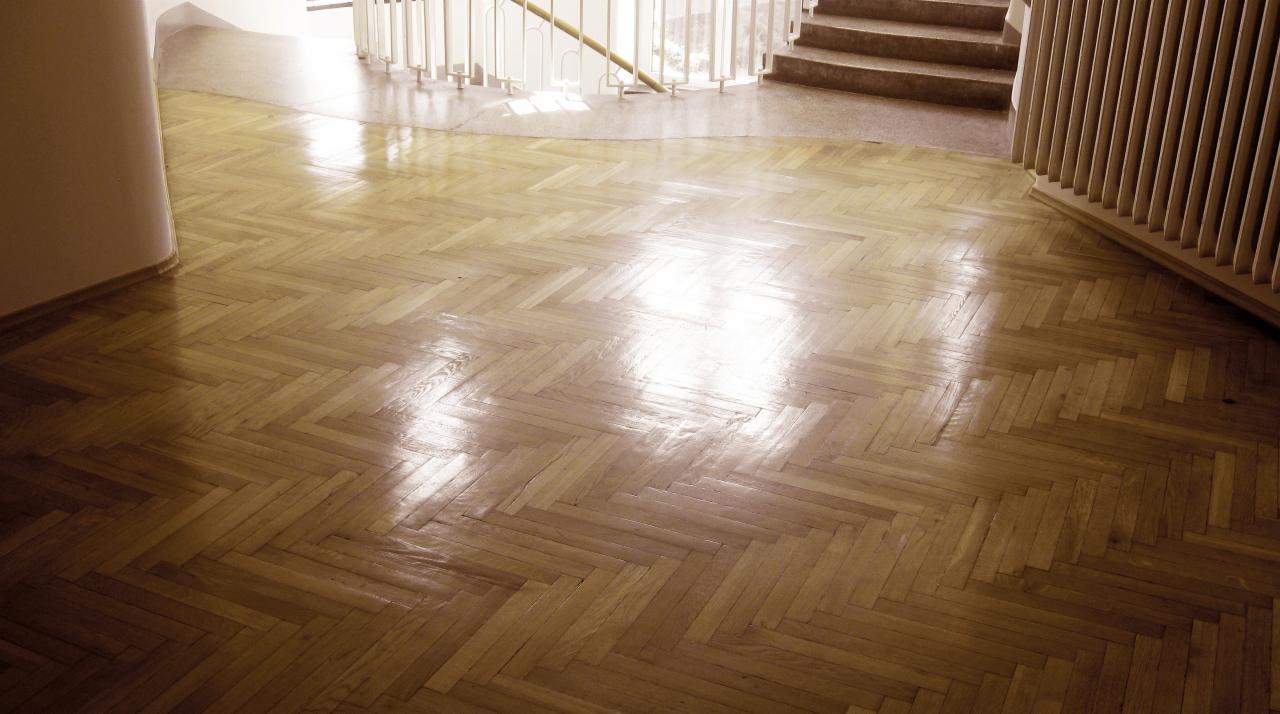 Błyszcząca, świeżo wycyklinowana podłoga parkietowa ułożona w jodełkę, widok na schody i białe balustrady w tle.