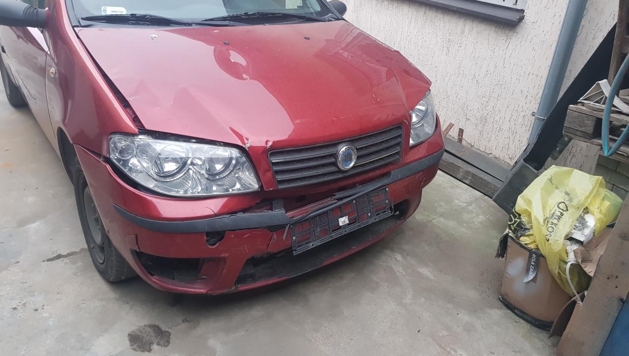Czerwony samochód osobowy Fiat Punto z uszkodzonym przodem, widoczne wgniecenia na masce i zniszczony zderzak, zaparkowany na betonowym podłożu przed budynkiem.