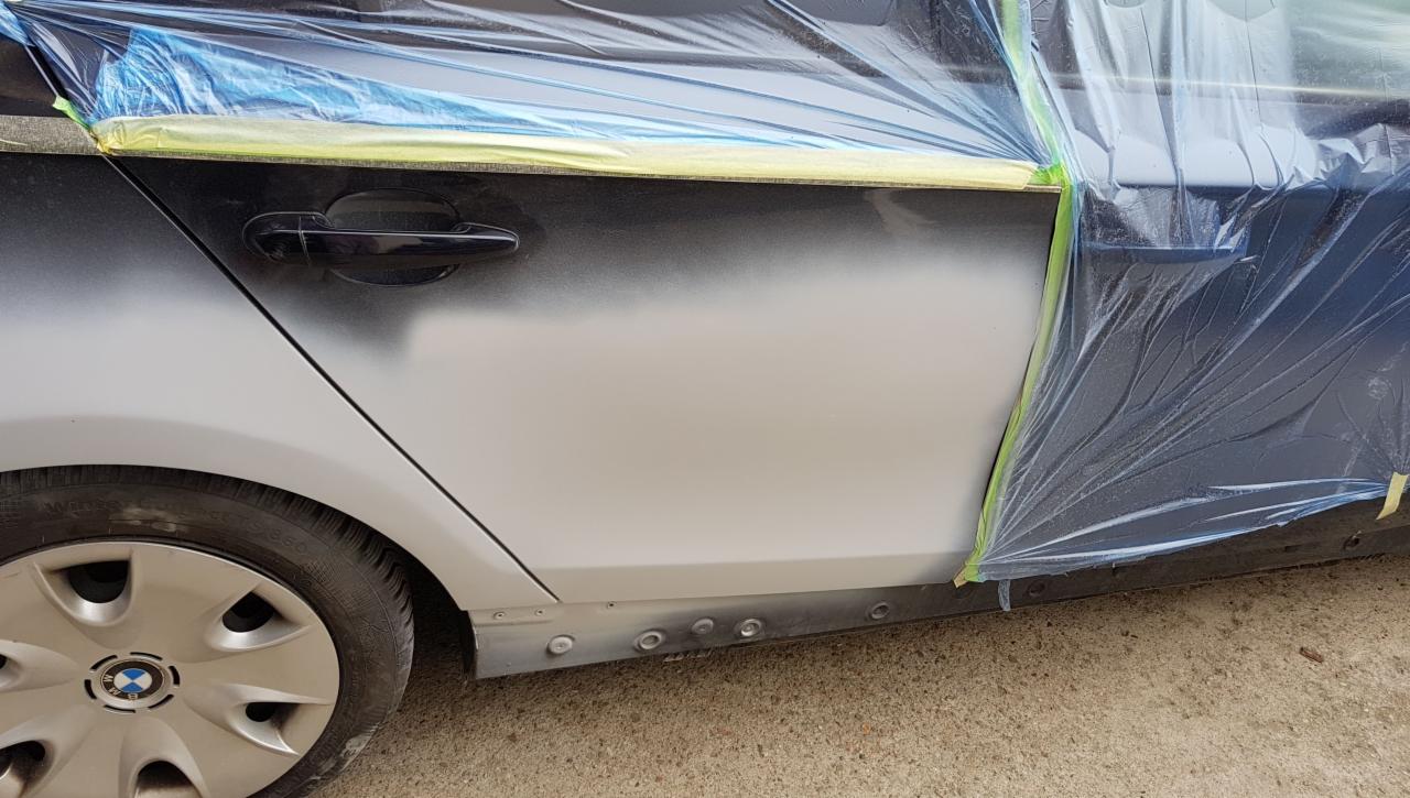 Samochód BMW w trakcie naprawy karoserii, częściowo zamaskowany folią i taśmą malarską, widoczne ślady szpachlowania i przygotowania do lakierowania.