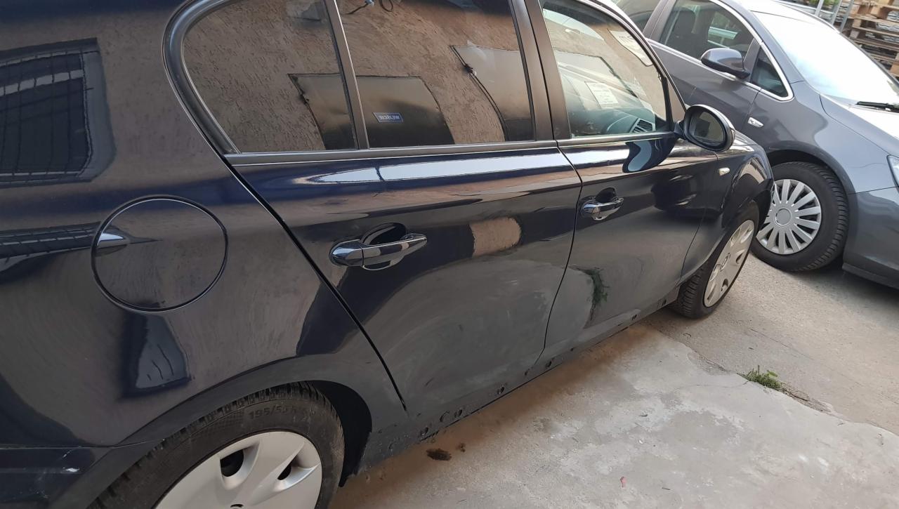 Bok ciemnego samochodu z widocznymi śladami napraw blacharskich i lakierniczych, zaparkowanego obok innego auta.