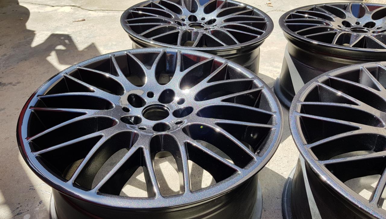 Cztery aluminiowe felgi samochodowe po renowacji, pomalowane na czarny kolor z metalicznym połyskiem, widoczne w ostrym świetle słonecznym.