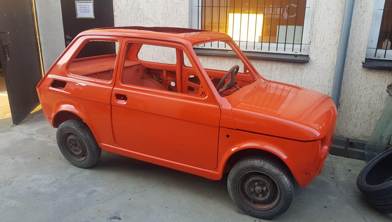 Pomarańczowy Fiat 126p bez szyb i wnętrza, po świeżym lakierowaniu, widoczny na tle warsztatu.