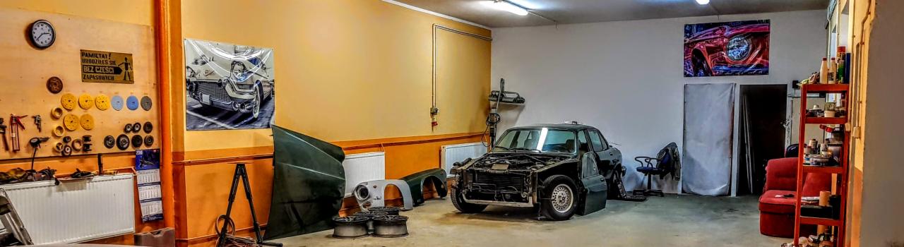 Warsztat samochodowy z rozmontowanym, klasycznym samochodem marki BMW, widoczne części karoserii obok, ściany z narzędziami i plakatami.