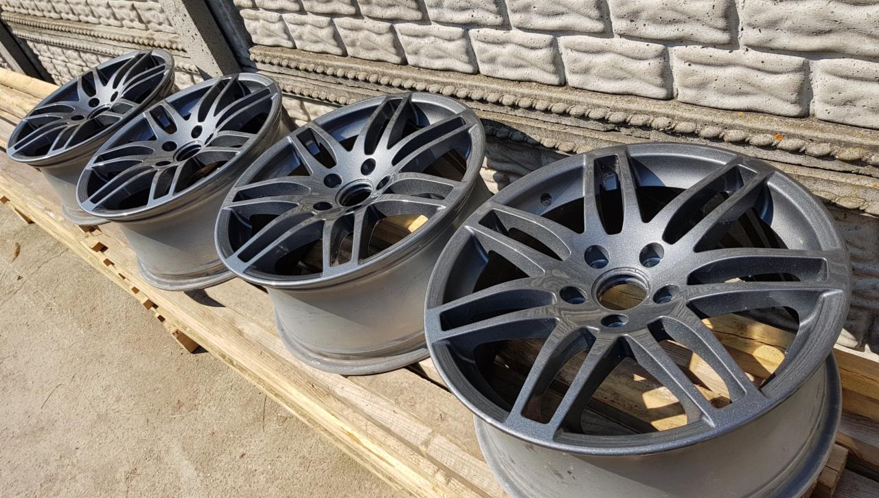 Cztery aluminiowe felgi samochodowe w kolorze grafitowym, leżące na drewnianej palecie na tle betonowego muru z dekoracyjnym gzymsem.