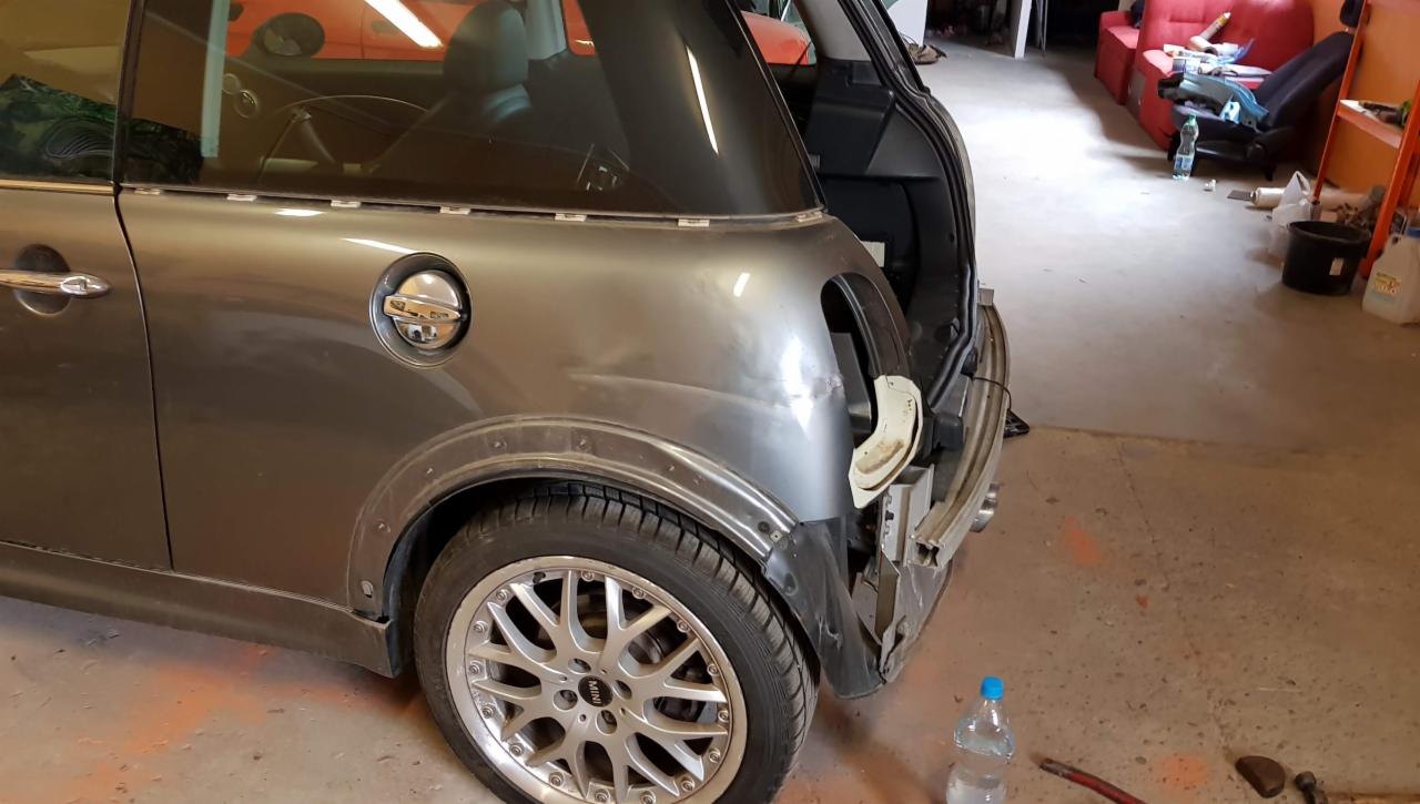 Szary Mini Cooper z uszkodzonym tylnym błotnikiem i zdemontowanym zderzakiem, widoczne ślady napraw blacharskich i przygotowania do lakierowania, na podłodze butelka wody i narzędzia.