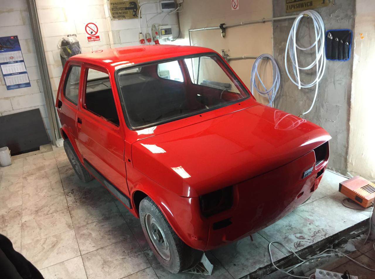 Czerwony samochód bez szyb i reflektorów, w trakcie prac renowacyjnych, stoi w warsztacie na podłodze wyłożonej płytkami.