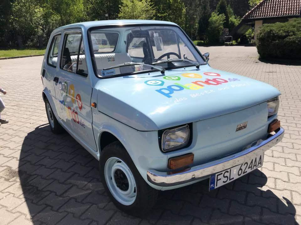Jasnoniebieski Fiat 126p z kolorowym nadrukiem 'polando.de' na masce, zaparkowany na brukowanym placu, widok z przodu pod lekkim kątem.