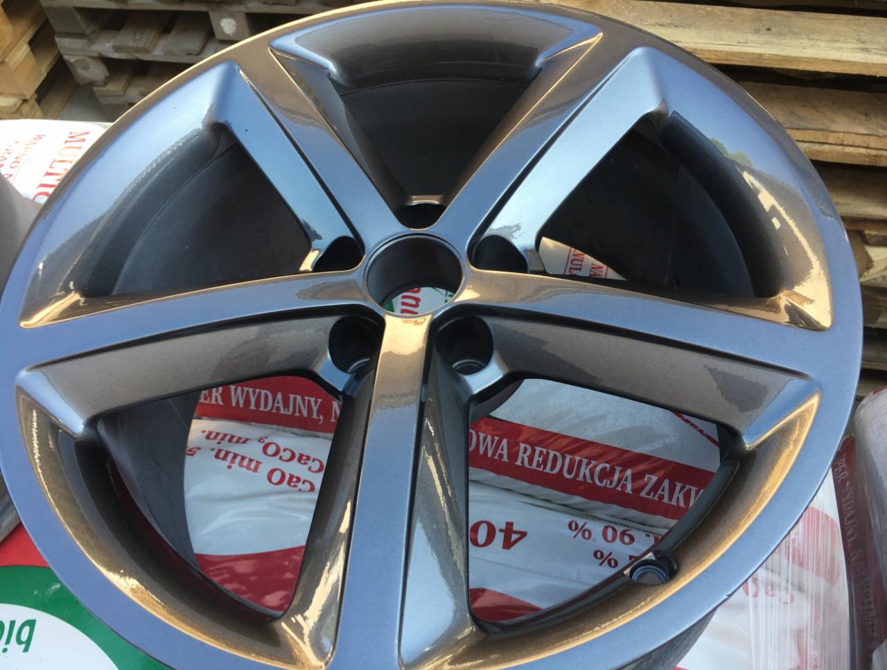 Szara felga aluminiowa z pięcioma ramionami, świeżo po renowacji, leżąca na palecie z workami zaprawy budowlanej w tle.