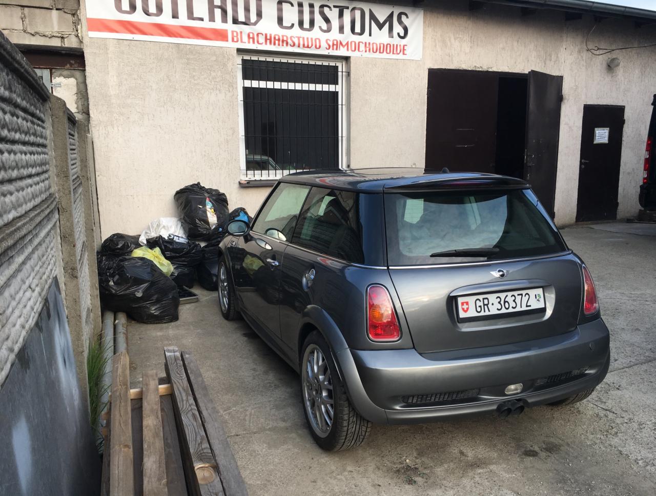 Szary Mini Cooper zaparkowany przed budynkiem warsztatu z szyldem 'OUTLAW CUSTOMS', widoczne worki ze śmieciami i drewniane deski.