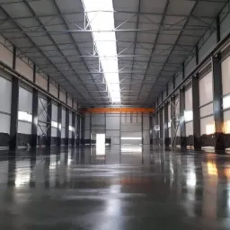 Błyszcząca, szara podłoga w hali przemysłowej z widocznymi odbiciami światła, pod suwnicą i stalową konstrukcją dachu.