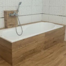 Biała wanna obudowana panelami imitującymi drewno, z zamontowaną chromowaną baterią z słuchawką prysznicową, na podłodze panele podłogowe w kolorze drewna, ściany wyłożone białymi płytkami...