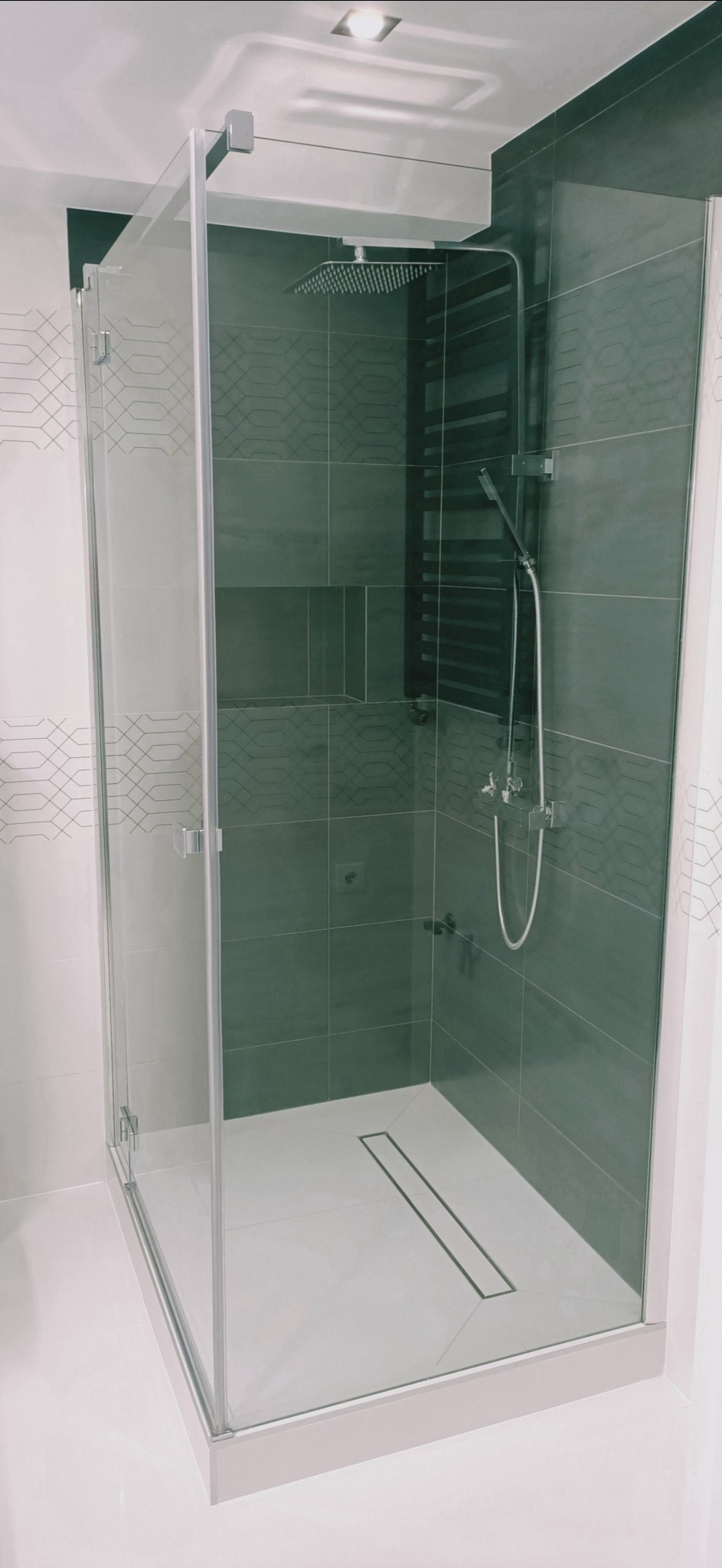 Nowoczesna kabina prysznicowa typu walk-in z deszczownicą i odpływem liniowym, wykończona szarymi płytkami z geometrycznym wzorem i białymi płytkami na zewnątrz.