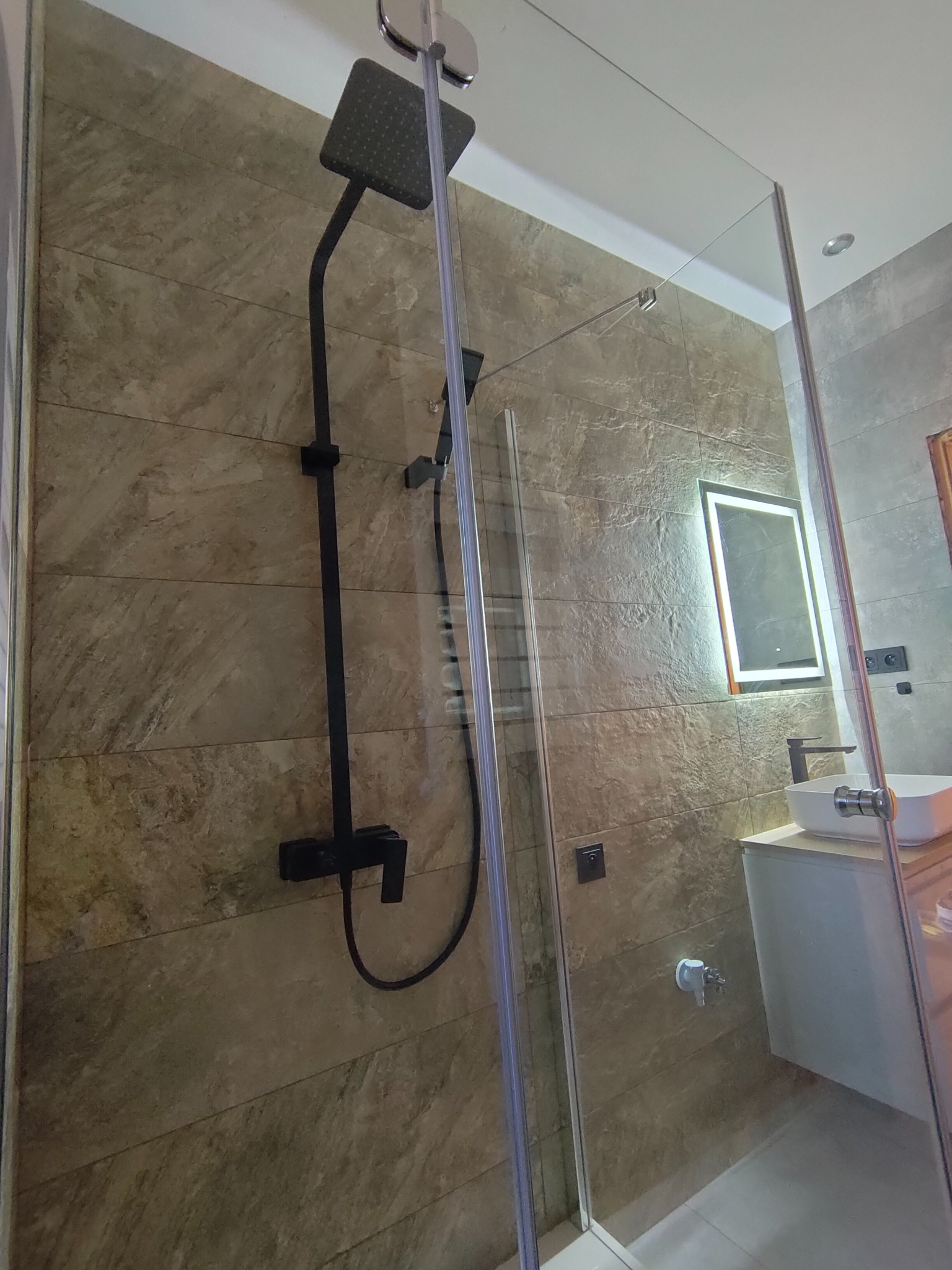 Nowoczesna kabina prysznicowa z czarną armaturą na tle płytek imitujących kamień, oświetlone lustro nad białą umywalką.