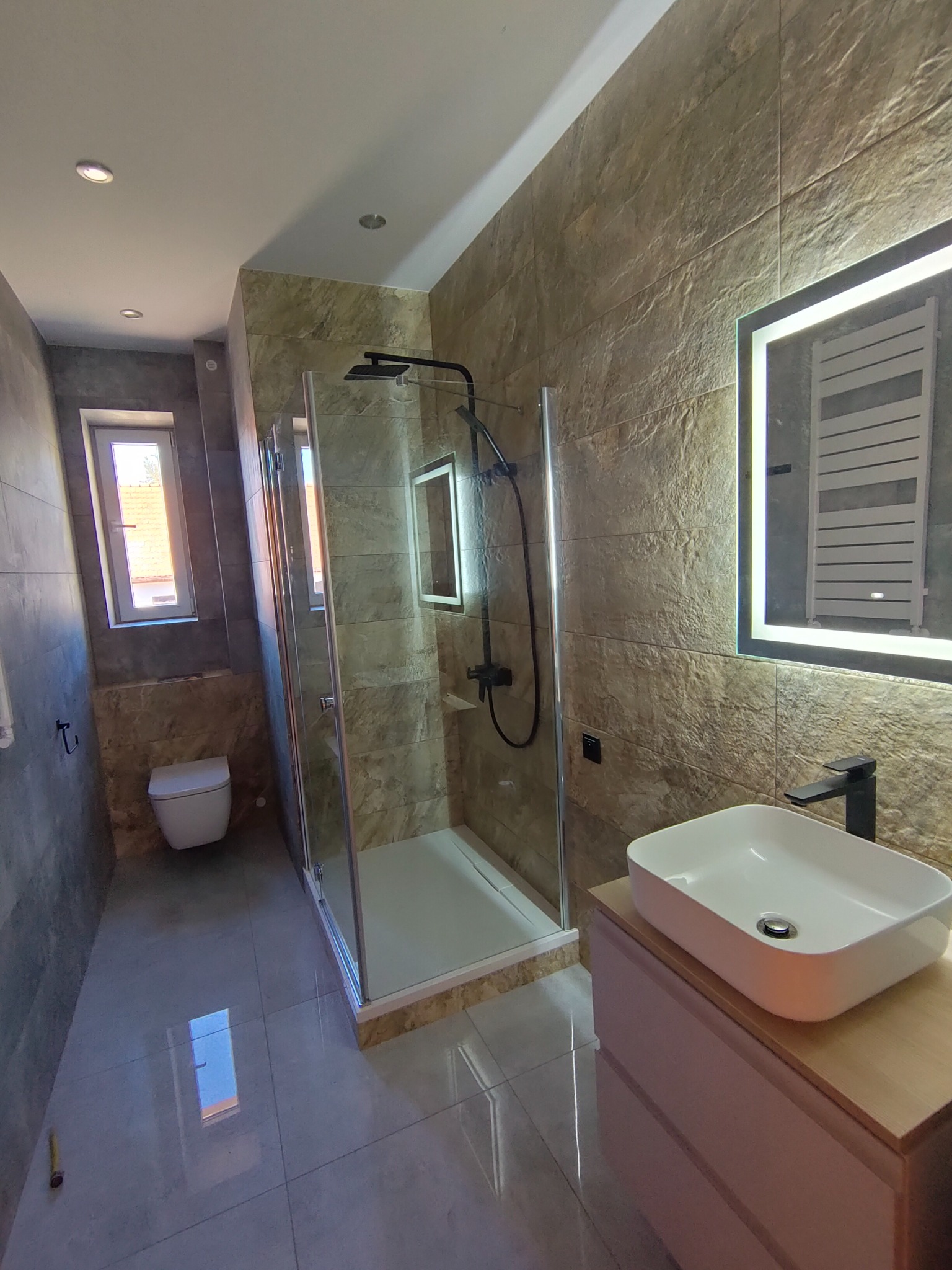 Nowoczesna łazienka z kabiną prysznicową typu walk-in, umywalką nablatową na szafce z drewnianym blatem, lustrem z podświetleniem LED i grzejnikiem drabinkowym, wykończona płytkami imitującymi...