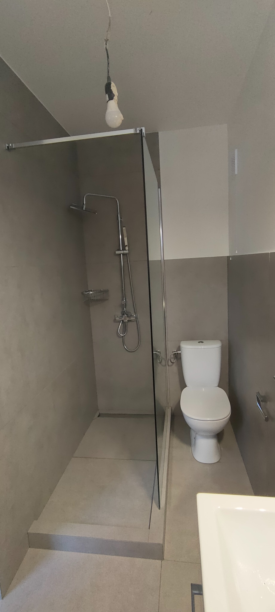 Nowoczesna łazienka z kabiną prysznicową typu walk-in z szarymi płytkami, białą toaletą i umywalką, surowa żarówka zwisająca z sufitu.