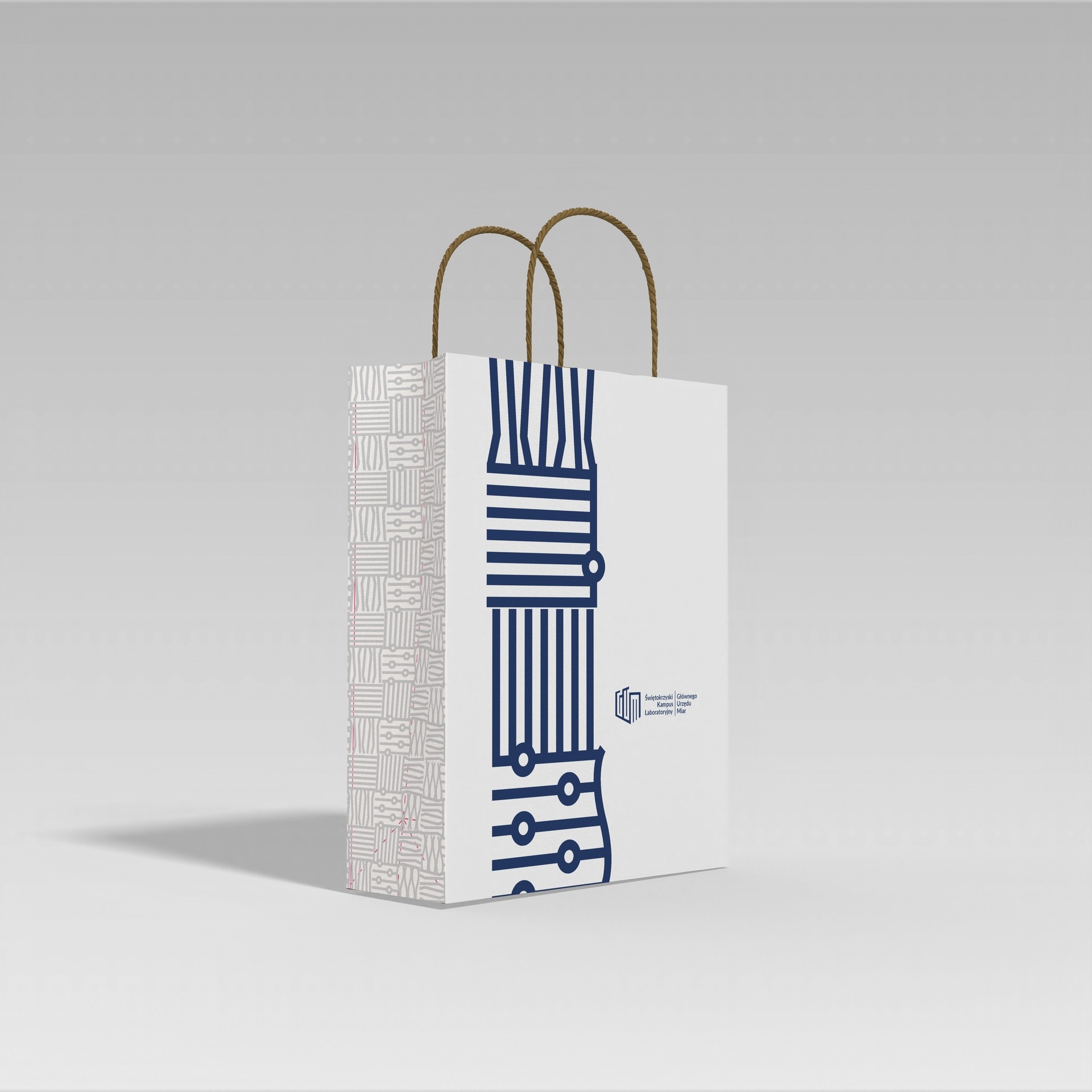 Biała torba papierowa z geometrycznym wzorem i logo uczelni, brązowe uchwyty sznurkowe, ustawiona na jasnoszarym tle.