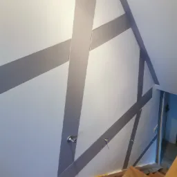 Ściana pomalowana w geometryczne, szare pasy w Elblągu, widoczne schody drewniane i zabezpieczenia podłogi folią malarską, detale wykończenia wnętrza.