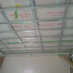 Stelaż sufitowy z profili metalowych, wypełniony materiałem izolacyjnym z nadrukiem, zabezpieczony zieloną taśmą, przewody zwisające z sufitu, fragment ściany z widoczną łatą.