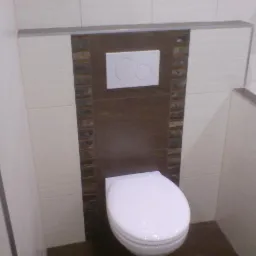 Zabudowany stelaż WC z białym przyciskiem spłukującym, obłożony brązowymi płytkami z mozaiką, komponujący się z jasnymi płytkami na ścianach.