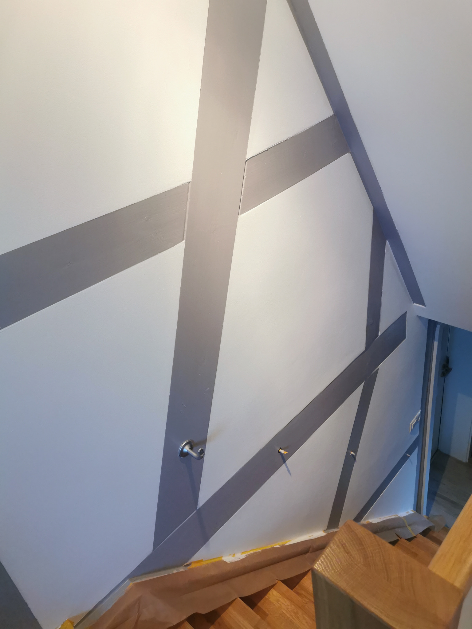 Ściana pomalowana w geometryczne, szare pasy w Elblągu, widoczne schody drewniane i zabezpieczenia podłogi folią malarską, detale wykończenia wnętrza.