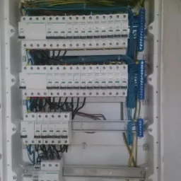 Rozdzielnia elektryczna z zainstalowanymi bezpiecznikami, listwami zaciskowymi i niebieską rurą karbowaną na dole obudowy, widoczne przewody w różnych kolorach.