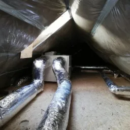 Centrala wentylacyjna z izolowanymi przewodami w przestrzeni poddasza, widoczne elementy montażowe i folia paroizolacyjna.