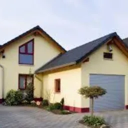 Żółta elewacja domu jednorodzinnego z garażem, widoczny fragment dachu z ciemnej dachówki, małe drzewko ozdobne przed garażem, kostka brukowa na podjeździe.