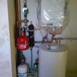 Nowy, jeszcze zapakowany piec centralnego ogrzewania z podłączonymi rurami miedzianymi, zbiornikiem wyrównawczym i białym zasobnikiem wody w kotłowni.