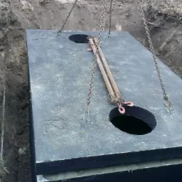 Wielkogabarytowy, betonowy zbiornik z otworami, opuszczany do wykopu za pomocą łańcuchów i zawiesi; widoczne rękawice robocze.