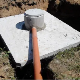 Betonowy zbiornik na szambo z pokrywą i podłączoną rurą PCV, częściowo zakopany w ziemi, widoczny wykop.