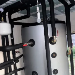 Szary zbiornik akumulacyjny z czarnymi, izolowanymi rurami i zaworami z czerwonymi uchwytami. Widoczne łączenia rur i manometr.