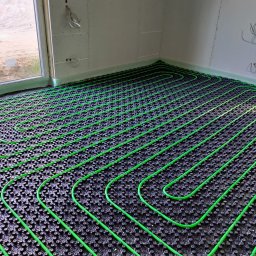 Termotechnika Piotr Skupiński - Zielone rury ogrzewania podłogowego ułożone na czarnej macie w surowym wnętrzu z oknem i widocznymi przewodami elektrycznymi.