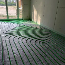 Zielone rury ogrzewania podłogowego ułożone spiralnie na szarej macie w jasnym pomieszczeniu z oknem i surowymi ścianami, przed wylaniem posadzki. Widok z góry.