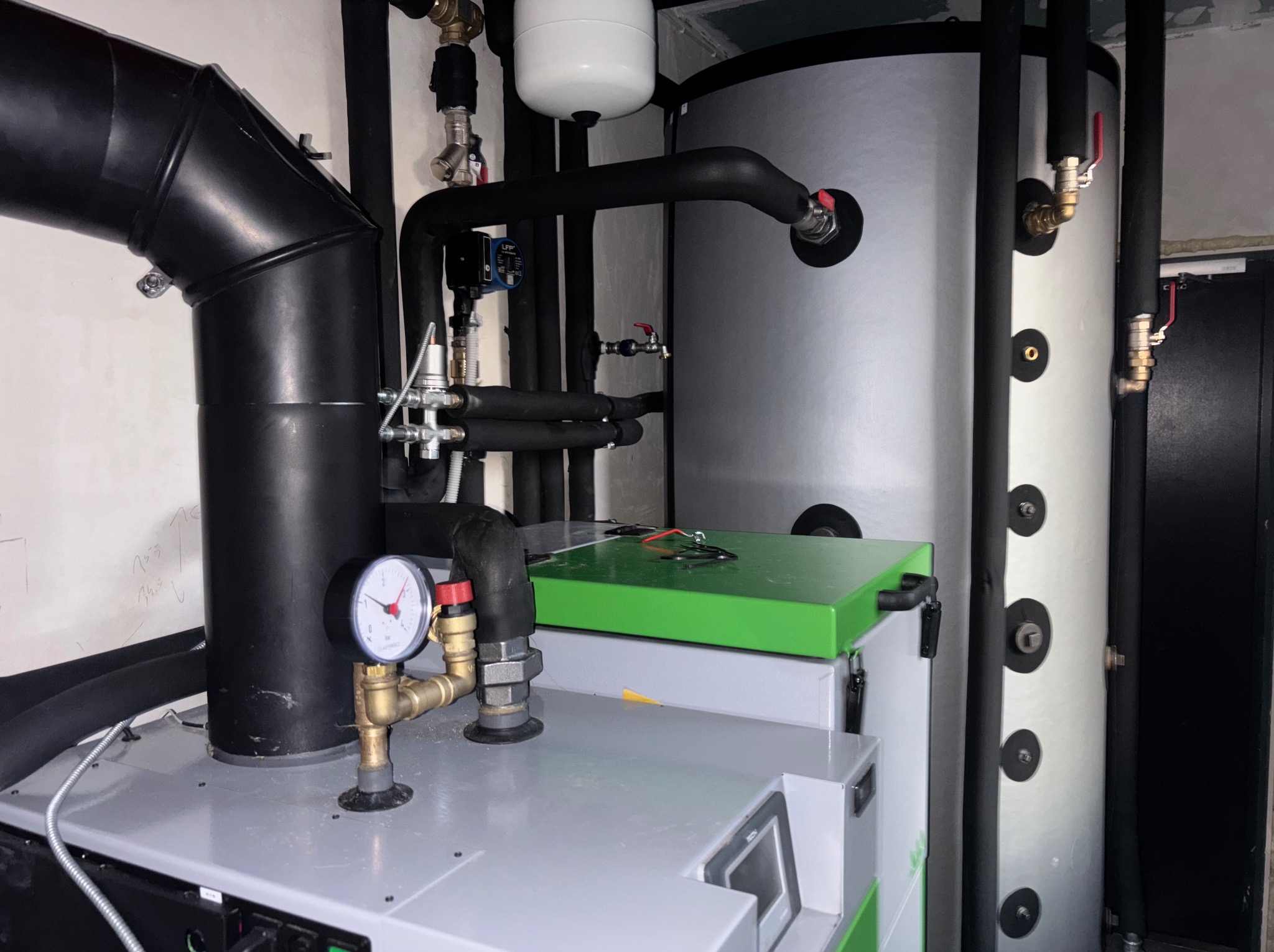 Nowoczesny piec CO z zielonym akcentem, widoczne rury, zawory i manometr. Instalacja grzewcza w kotłowni, srebrny zbiornik buforowy. Elementy instalacji w izolacji.