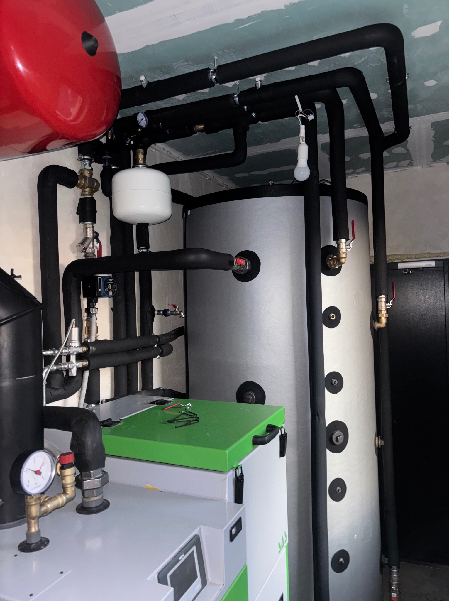 Nowoczesna kotłownia: srebrne zbiorniki, czarne rury z izolacją, czerwony zasobnik, manometry i zawory. Widok na kompleksowy system grzewczy w pomieszczeniu.