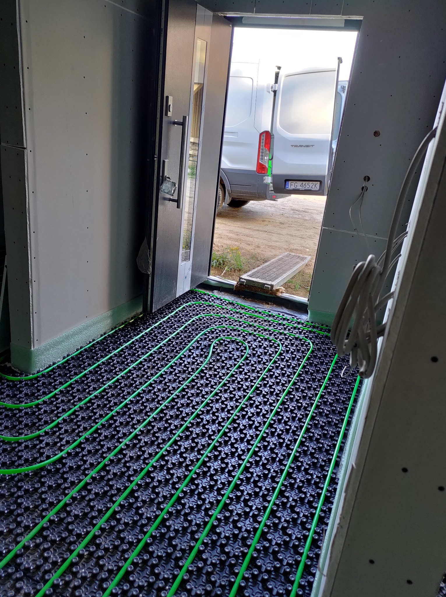 Zielone rury ogrzewania podłogowego ułożone na czarnej macie w pomieszczeniu z widokiem na samochód dostawczy przez otwarte drzwi.