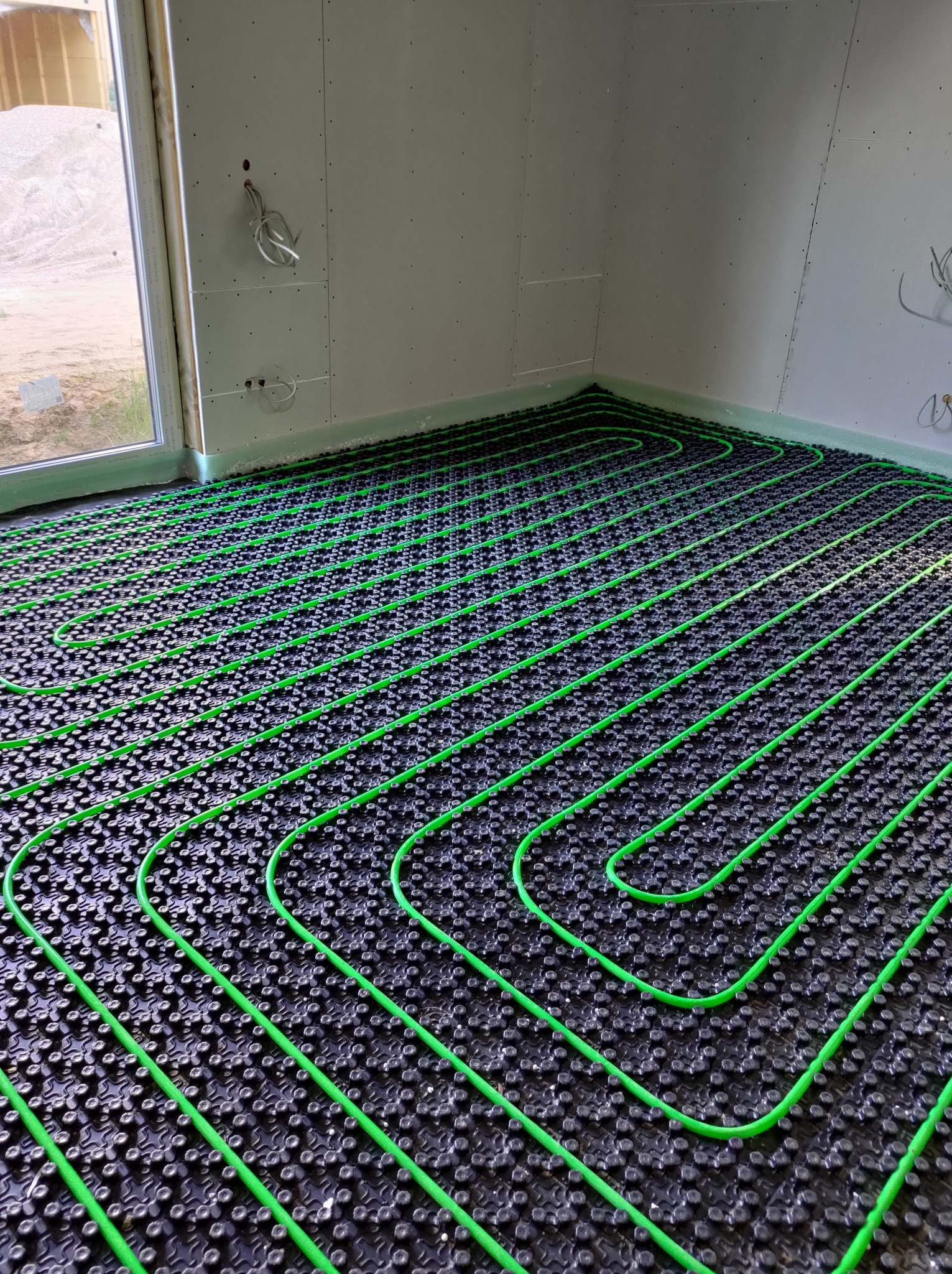 Zielone rury ogrzewania podłogowego ułożone na czarnej macie w surowym wnętrzu z oknem i widocznymi przewodami elektrycznymi.