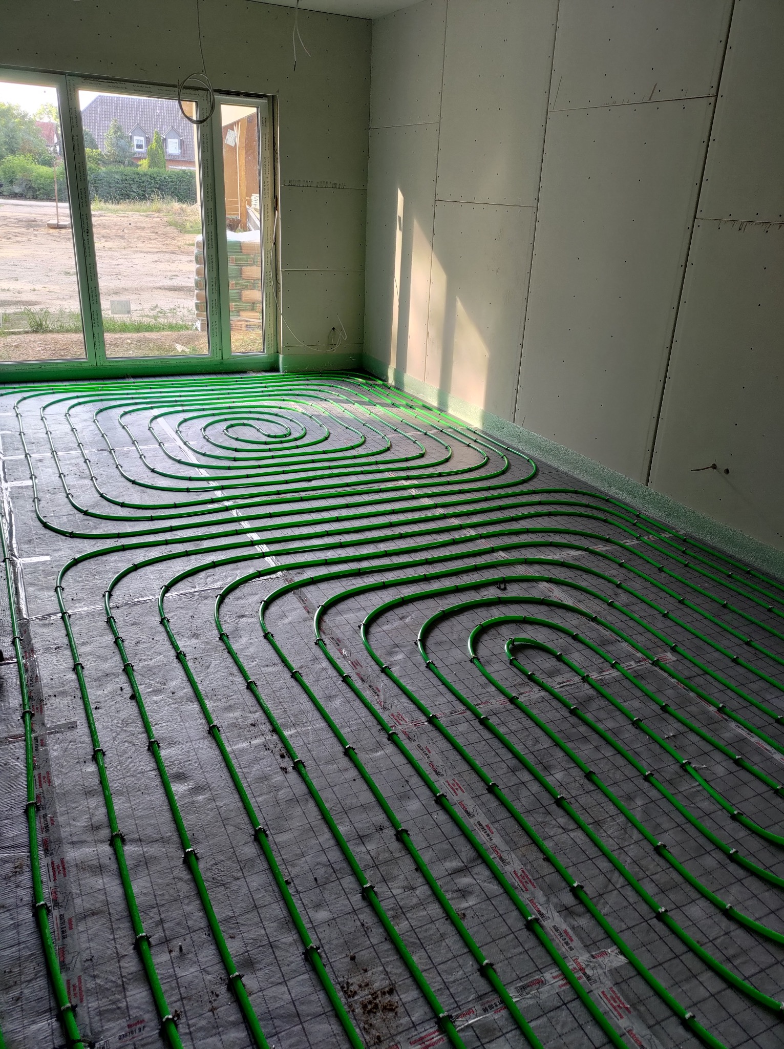 Zielone rury ogrzewania podłogowego ułożone spiralnie na szarej macie w jasnym pomieszczeniu z oknem i surowymi ścianami, przed wylaniem posadzki. Widok z góry.