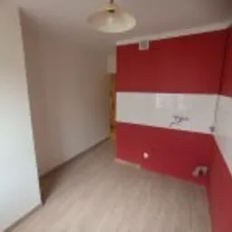 Puste pomieszczenie po remoncie z panelami podłogowymi, ściana pomalowana częściowo na czerwono z białymi płytkami w górnej części, widoczna instalacja wodna i elektryczna, sufit z lampą.