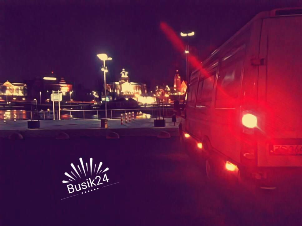 Biały bus z włączonymi światłami stopu na tle nocnego pejzażu miasta z oświetlonymi budynkami, logo 'Busik24' w lewym dolnym rogu.